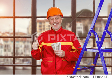 Portrait of smiling elder man with dollar baknotes showing like gesture. Handyman construction worker with his salary standing indoors. 114034619