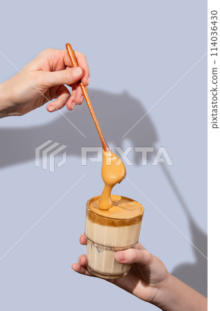 Dalgona coffee. One hand holds a glass of iced coffee and milk, the other hand puts whipped foam  114036430