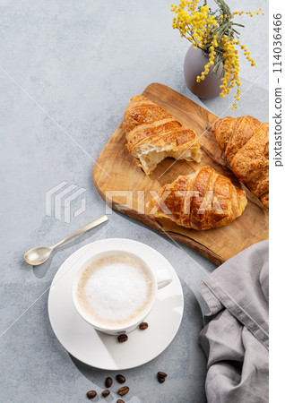 A cup of aromatic cappuccino coffee and fresh croissants on a cutting board on a blue background A cup of aromatic cappuccino coffee and fresh croissants on a cutting board on a blue background 114036466