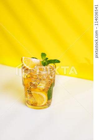 A glass of iced tea with lemon and mint on a yellow and white background with a shadow. 114036541