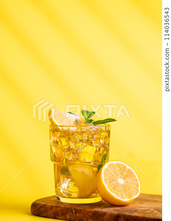 A glass of iced tea with lemon and mint on a wooden board on a yellow background with a shadow. A glass of iced tea with lemon and mint on a wooden board on a yellow background with a shadow. 114036543