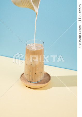 Iced coffee in a tall glass with a milk jug pouring milk on a blue and yellow background with shadow Iced coffee in a tall glass with a milk jug pouring milk on a blue and yellow background with shadow 114036629