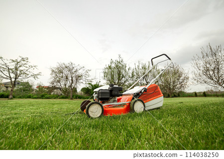 side view of modern orange-grey gasoline lawn mower cutting bright lush green grass. Gardening work tools. Rotary lawn mower machine on lawn. Professional lawn care service. Place for text 114038250