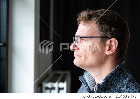 Indoor portrait of a 45 yo white man looking through his window at home, Brussels, Belgium Indoor portrait of a 45 yo white man looking through his window at home, Brussels, Belgium 114038455
