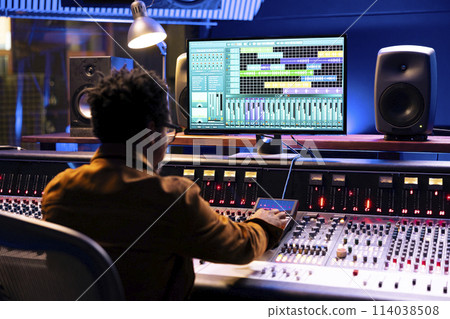 African american audio expert operating editing software to adjust sound settings, control room with mixing console in studio. Professional technician creating new tracks for an album. 114038508