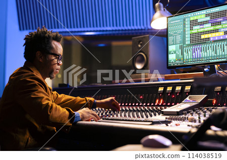 African american sound engineer works on mixing console with sliders and knobs, looking at digital software to record and process tunes. Audio technician creating new music, control room board. African american sound engineer works on mixing console with sliders and knobs, looking at digital software to record and process tunes. Audio technician creating new music, control room board. 114038519