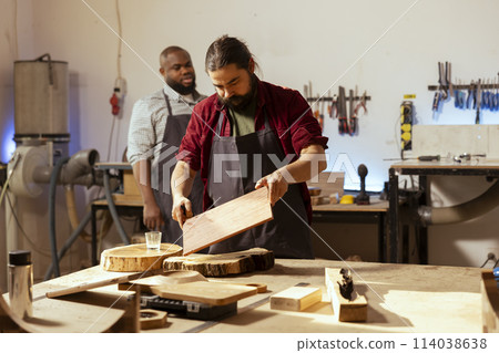 Craftsperson at workbench starting restoration project on wood block next to african american coworker. Carpenter preparing to color plank with paintbrush near apprentice solving tasks behind Craftsperson at workbench starting restoration project on wood block next to african american coworker. Carpenter preparing to color plank with paintbrush near apprentice solving tasks behind 114038638