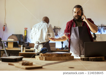 Smiling manufacturer discussing on telephone call with client inquiring about furniture assembling commission progress. Cheerful carpenter in assembly shop talking on mobile phone with customer Smiling manufacturer discussing on telephone call with client inquiring about furniture assembling commission progress. Cheerful carpenter in assembly shop talking on mobile phone with customer 114038645