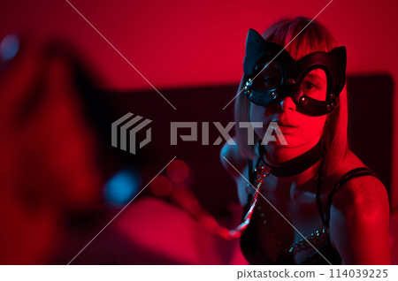 Blonde girl in a leather mask and handcuffs in the bedroom in red blue neon light. Role-playing games. Blonde girl in a leather mask and handcuffs in the bedroom in red blue neon light. Role-playing games. 114039225