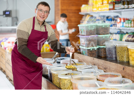 Man in supermarket, scooping marinated olives from bucket with ladle Man in supermarket, scooping marinated olives from bucket with ladle 114042846