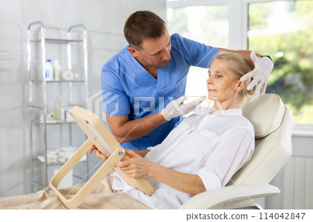 Senior woman looking at mirror while cosmetologist explaining facial procedure 114042847