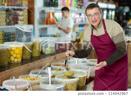 Seller man in supermarket showing and scooping marinated green olives 114042967