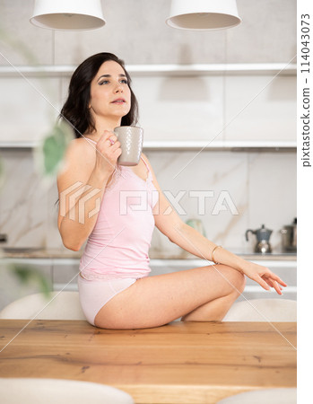Woman in white lady singlet lingerie and underpants is sitting on kitchen table 114043073