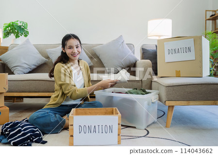 Asian young woman packing clothes at home, putting on stuff into donate box with second hand clothes Asian young woman packing clothes at home, putting on stuff into donate box with second hand clothes 114043662