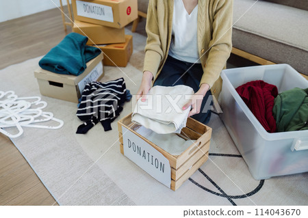 Asian young woman packing clothes at home, putting on stuff into donate box with second hand clothes 114043670