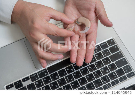 Close-up of a man's hands with a hearing aid near a laptop. Close-up of a man's hands with a hearing aid near a laptop. 114045945