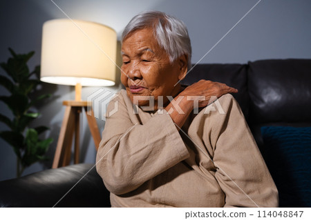 senior woman suffering from neck and shoulder pain while sitting on sofa in living room at night 114048847