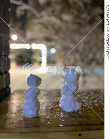 Two mini snowmen made of snowballs stand on a wooden table outside 114048878