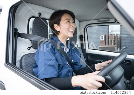 A middle-aged woman in work clothes driving a truck. Image of the transportation and delivery industry. 114052894