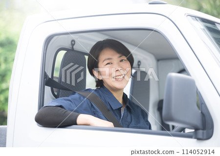 A middle-aged woman in work clothes driving a truck, looking at the camera, an image of the transportation and delivery industry 114052914