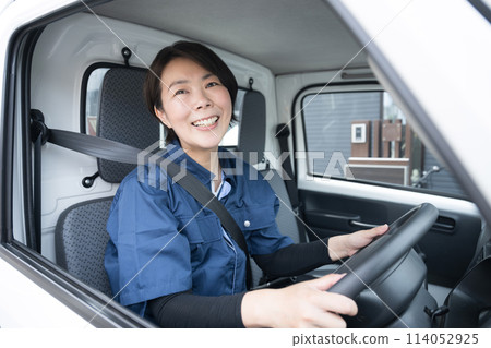 A middle-aged woman in work clothes driving a truck, looking at the camera, image of transportation and delivery industry A middle-aged woman in work clothes driving a truck, looking at the camera, image of transportation and delivery industry 114052925