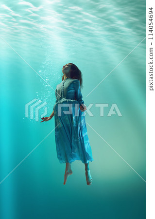 Rebirth. Tender young redhead girl in elegant dress diving into blue water, swimming in direction to sunlight Rebirth. Tender young redhead girl in elegant dress diving into blue water, swimming in direction to sunlight 114054694