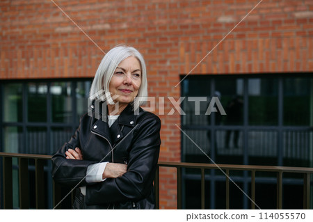Portrait of stylish mature woman with gray hair on city street. Older woman in leather jacket with soft smile. 114055570