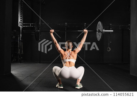 Rear view of woman performing overhead squat pressing barbell up, standing up. Routine workout for physical and mental health. Rear view of woman performing overhead squat pressing barbell up, standing up. Routine workout for physical and mental health. 114055621