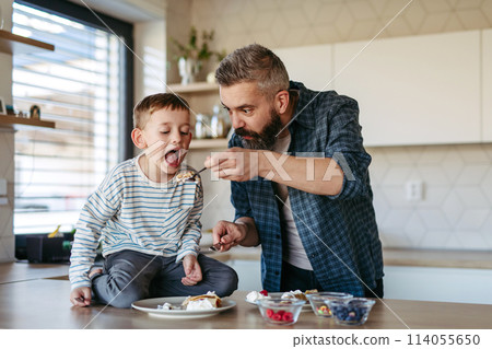 Boy eating pancake with fruit, sweets. Father spending time with son at home, making snack together, cooking. Fathers day concept. 114055650