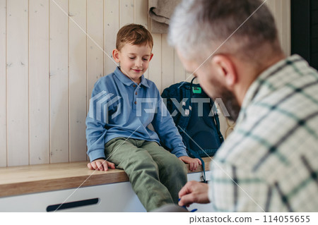 Father helping son get ready for kindergarten, preschool. Putting coat, shoes on, handing lunchbox with snacks. Father helping son get ready for kindergarten, preschool. Putting coat, shoes on, handing lunchbox with snacks. 114055655
