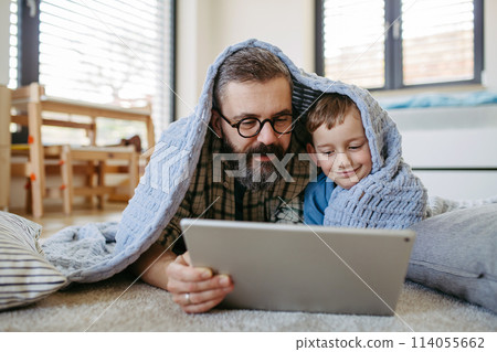 Little boy watching cartoon movie on tablet with father, lying under blanket on floor in kids room. Dad explaining technology to son, digital literacy for kids. Little boy watching cartoon movie on tablet with father, lying under blanket on floor in kids room. Dad explaining technology to son, digital literacy for kids. 114055662