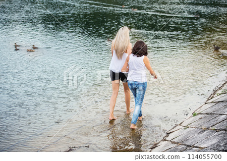 Adult daughter spending time with her mother. Mom and daughter outdoors, walking barefoot in water of lake reservoir, lake embankment. Unconditional, deep maternal love, Mother's Day. 114055700