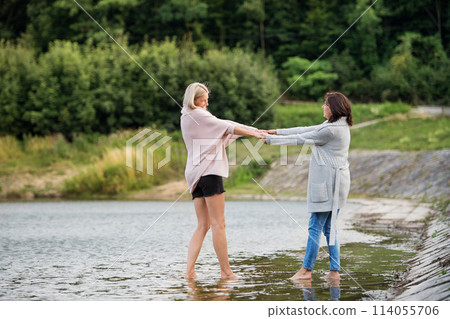 Adult daughter spending time with her mother. Mom and daughter outdoors, walking barefoot in water of lake reservoir, lake embankment. Unconditional, deep maternal love, Mother's Day. 114055706