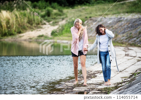 Adult daughter spending time with her mother. Mom and daughter outdoors, walking barefoot in water of lake reservoir, lake embankment. Unconditional, deep maternal love, Mother's Day. 114055714