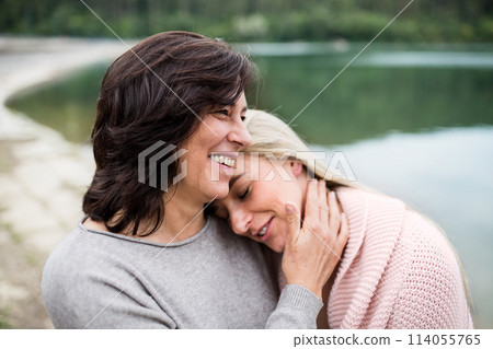 Adult daughter spending time with her mother. Mom and daughter embracing outdoors, standing by reservoir, lake embankment. Unconditional, deep maternal love, Mother's Day concept. Adult daughter spending time with her mother. Mom and daughter embracing outdoors, standing by reservoir, lake embankment. Unconditional, deep maternal love, Mother's Day concept. 114055765