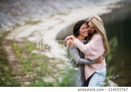 Adult daughter spending time with her mother. Mom and daughter embracing outdoors, standing by reservoir, lake embankment. Unconditional, deep maternal love, Mother's Day concept. Adult daughter spending time with her mother. Mom and daughter embracing outdoors, standing by reservoir, lake embankment. Unconditional, deep maternal love, Mother's Day concept. 114055767