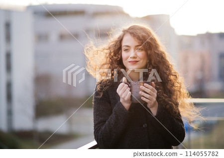 Beautiful woman with curly hair standing on terrace, drinking coffee during cold spring day. Morning sun, hair blowing in wind. 114055782