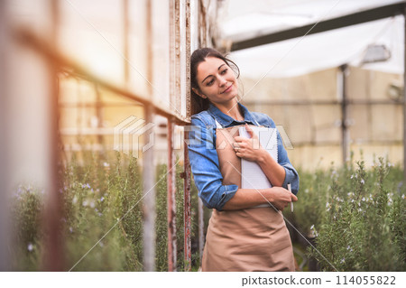 Small greenhouse business. Businesswoman selling flowers and seedlings, standing in the middle of plants, enjoying beautiful crops, smiling. 114055822