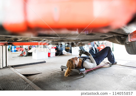 Female auto mechanic lying on mechanic creeper under car, inspecting and repairing vehicle. Beautiful woman working in a garage, wearing blue coveralls. 114055857