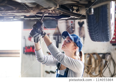 Female auto mechanic elevating car on car lift, working underneath. Beautiful woman working in a garage, wearing blue coveralls. Female auto mechanic elevating car on car lift, working underneath. Beautiful woman working in a garage, wearing blue coveralls. 114055864
