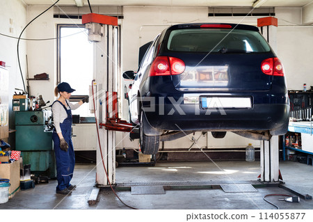 Female auto mechanic elevating car on car lift, working underneath. Beautiful woman working in a garage, wearing blue coveralls. 114055877