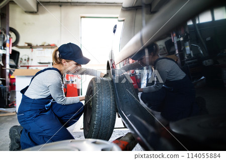 Female auto mechanic changing tieres in auto service. Beautiful woman kneeling by wheel in a garage, wearing blue coveralls. 114055884