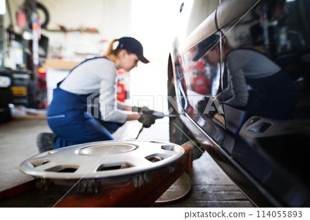 Female auto mechanic changing tieres in auto service. Beautiful woman kneeling by wheel in a garage, wearing blue coveralls. Female auto mechanic changing tieres in auto service. Beautiful woman kneeling by wheel in a garage, wearing blue coveralls. 114055893