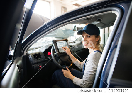 Female auto mechanic using diagnostic tool, scanner, running diagnostic on car. Beautiful woman sitting inside of car, performing diagnostic test. Female auto mechanic using diagnostic tool, scanner, running diagnostic on car. Beautiful woman sitting inside of car, performing diagnostic test. 114055901