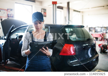 Female auto mechanic using diagnostic tool, scanner, running diagnostic on car. Beautiful woman working in a garage, wearing blue coveralls. 114055925