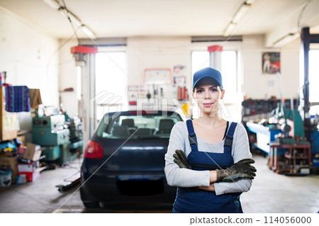 Female auto mechanic repairing, maintaining car. Beautiful woman standing in a garage, wearing blue coveralls. 114056000
