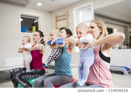 Group exercise class, mother working out with baby on fitball in gym. Moms staying active while boding with babies. 114056016