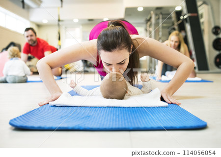 Mother smiling at baby while performing push-ups, kissing infant. Group exercise class, fitness or pilates for parents. Moms staying active while boding with babies. 114056054