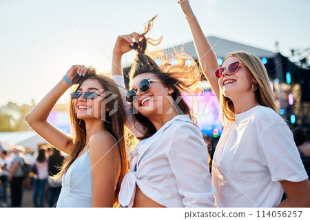 Girls in sunglasses enjoy music at sunny beach fest. Friends dance, laugh with stage behind. Casual summer wear, youth culture embraced at seaside concert. Holiday vibe as trio celebrates friendship. 114056257