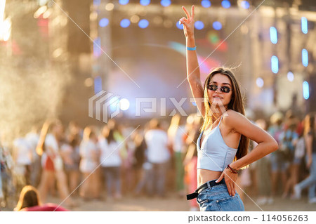 Happy young woman enjoys summer music festival, raises peace sign with sunlit crowd and stage background. Cheerful festivalgoer in stylish sunglasses dances at beach event, vibrant celebration mood. Happy young woman enjoys summer music festival, raises peace sign with sunlit crowd and stage background. Cheerful festivalgoer in stylish sunglasses dances at beach event, vibrant celebration mood. 114056263